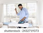 Cheerful young Black man enjoying house work, ironing white fabric towel, dancing to music at board, enjoying domestic activities, laundry, housework at home, having fun