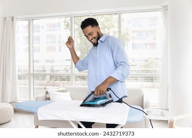 Cheerful young Black man enjoying house work, ironing white fabric towel, dancing to music at board, enjoying domestic activities, laundry, housework at home, having fun - Powered by Shutterstock