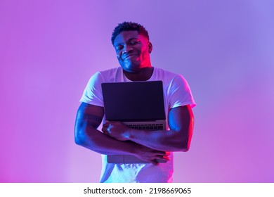 Cheerful Young Black Man Hugging Laptop With Empty Screen In Neon Light, Mockup For Website. Tech Nerd Embracing Portable Pc, Offering Copy Space For Your Ad. Technology Lover Concept