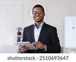 Cheerful young black man manager in glasses holding digital tablet and smiling at camera, office interior, copy space. African american businessman using business application on pad