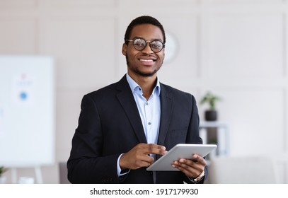 Cheerful young black man manager in glasses holding digital tablet and smiling at camera, office interior, copy space. African american businessman using business application on pad