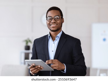 Cheerful young black man manager in glasses holding digital tablet and smiling at camera, office interior, copy space. African american businessman using business application on pad - Powered by Shutterstock