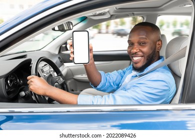 Cheerful young black man sitting in luxury car, showing cellphone with white blank screen, recommending newest mobile application for navigation, mockup. Automobile, driving and modern technologies