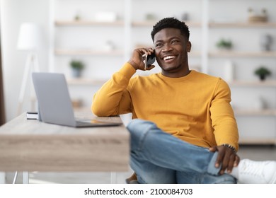 Cheerful Young Black Man Sitting At Worktable With Laptop, Looking For Job On Internet At Home, Talking On Phone With Employer Or HR Representative, Looking At Copy Space And Smiling