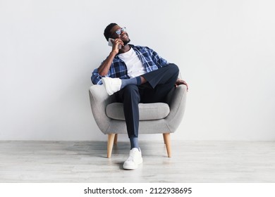 Cheerful Young Black Man Speaking On Cellphone, Sitting In Armchair, Having Phone Conversation With Friend Against White Studio Wall. Millennial African American Guy Communicating Online On Smartphone