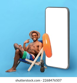 Cheerful young black man in swimwear sitting in lounge chair, pointing at big cellphone with blank screen, recommending new mobile app, showing thumb up on blue studio background, mockup, collage - Powered by Shutterstock