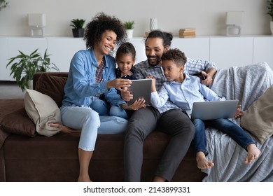 Cheerful young Black parents and curious little gen Z boy and girl resting together on home sofa, using laptop, tablet computer gadget, shopping internet, chatting online on social media - Powered by Shutterstock