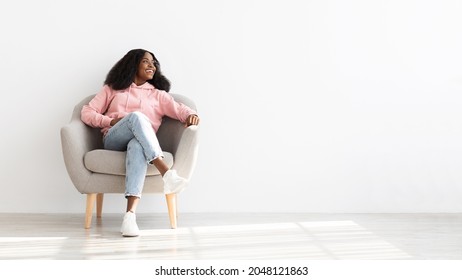 Cheerful Young Black Woman With Bushy Hair In Casual Outfit Sitting In Arm Chair At Home, Looking At Copy Space And Smiling. Pretty Happy Afro American Lady Spending Weekend Alone, Panorama