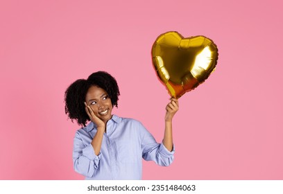 Cheerful young black woman in casual outfit look at inflatable gold heart balloon, isolated on pink studio background. Ad, offer, party and love, dream - Shutterstock ID 2351484063