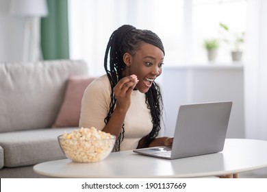 Cheerful young black woman with dreadlocks watching comedy movie on laptop at home, sitting on floor by coffee table, eating popcorn and laughing. African american lady having fun during isolation