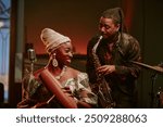 Cheerful young Black woman and man playing guiro and saxophone in jazz band on stage in local bar