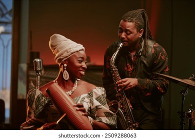 Cheerful young Black woman and man playing guiro and saxophone in jazz band on stage in local bar