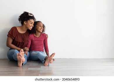 Cheerful Young Black Woman Mother And Teen Girl Daughter In Casual Sitting On Floor, Hugging And Looking At Copy Space For Advertisement Or Text, White Empty Wall Background, Panorama