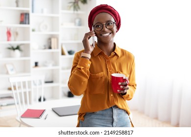 Cheerful young black woman with red headscarf and eyeglasses talking on phone with lover or girlfriend while having coffee break at office, holding red mug, looking at copy space and smiling, panorama - Powered by Shutterstock