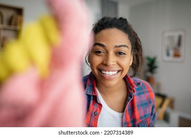 Cheerful young black woman in rubber gloves washes mirror with rag, enjoys cleaning in living room interior, close up, copy space. Perfectionism, housework, cleanliness and hygiene at home, lifestyle