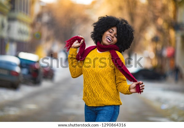 Cheerful young black woman in scarf and\
sweater is smiling to the city street.\
Closeup.
