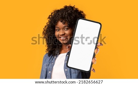 Cheerful young black woman showing modern cell phone with white empty screen on yellow studio background, panorama, copy space. Positive african american pretty lady recommending mobile app, collage