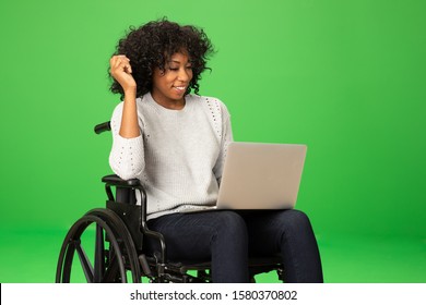 Cheerful Young Black Woman Sits In Wheelchair, Using Laptop. Portrait Of Disabled Woman Using Computer On Greenscreen