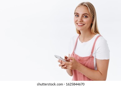 Cheerful Young Blond Female In Pink Overalls, T-shirt Standing Half-turned And Looking Camera With Happy Smile, Holding Smartphone, Messaging Or Using Social Media When Someone Called Her Name