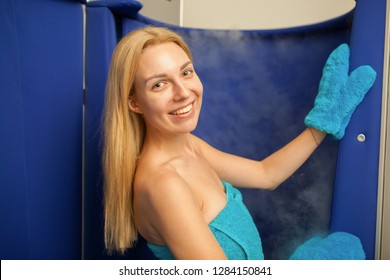 Cheerful young blond haired woman smiling to the camera over her shoulder, entering cryotheraby sauna booth. Attractive woman going for full body cryotherapy procedure at beauty salon