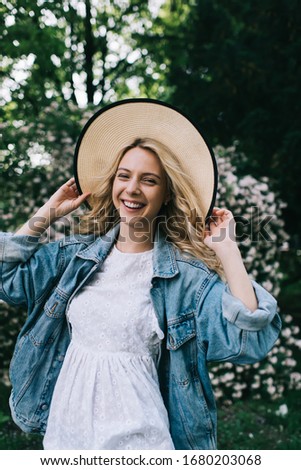 Cheerful young blonde woman in hat and jeans jacket looking at camera and laughing happily while standing in blooming park during summertime