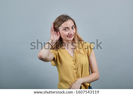 Cheerful young blonde woman keeping palm on ear, listening, smiling wearing fashion yellow shirt isolated on gray background in studio. People sincere emotions, lifestyle concept.