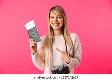Cheerful young blonde woman wearing sweater standing isolated over pink background, showing passport with flight tickets