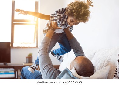 Cheerful Young Boy Having Fun With His Father On Sofa. Little African Son Playing With Father On Couch With Open Outstretched Arms. Black Child Enjoying Playing With Dad At Home.