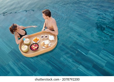 Cheerful young boyfriend and girlfriend standing in swimming pool, eating delicious breakfast and chatting - Powered by Shutterstock