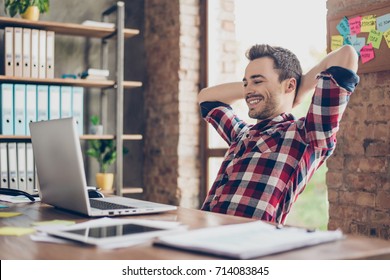 Cheerful Young Brunet Guy Is Watching At His Laptop Screen, At His Work Place, With Arms Behind The Head, Resting, Smiling, In The Office