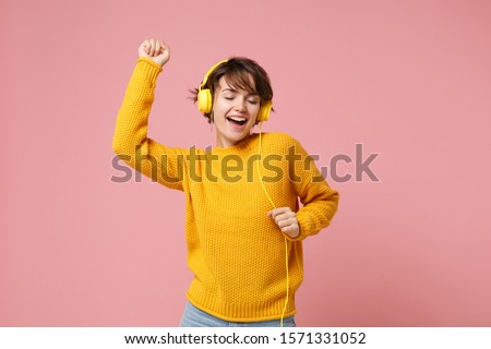Cheerful young brunette woman girl in yellow sweater posing isolated on pastel pink wall background studio portait. People lifestyle concept. Mock up copy space. Listen music with headphones, dancing