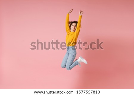 Cheerful young brunette woman girl in yellow sweater posing isolated on pastel pink background in studio. People lifestyle concept. Mock up copy space. Having fun fooling around rising hands, jumping