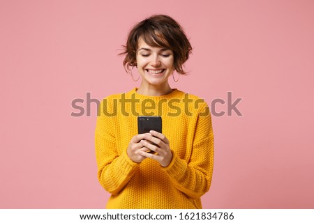 Cheerful young brunette woman girl in yellow sweater posing isolated on pastel pink wall background studio portait. People lifestyle concept. Mock up copy space. Using mobile phone typing sms message