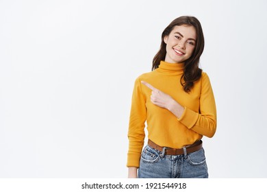 Cheerful young brunette woman smiling, pointing finger left at logo, showing copy space for your advertisement, standing in yellow sweater on white background. - Powered by Shutterstock