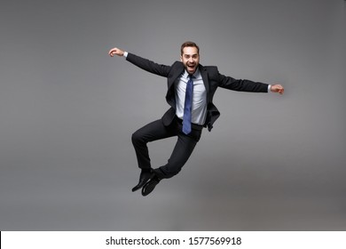 Cheerful Young Business Man In Classic Black Suit Shirt Tie Posing Isolated On Grey Background. Achievement Career Wealth Business Concept. Mock Up Copy Space. Jumping, Spreading Hands, Having Fun