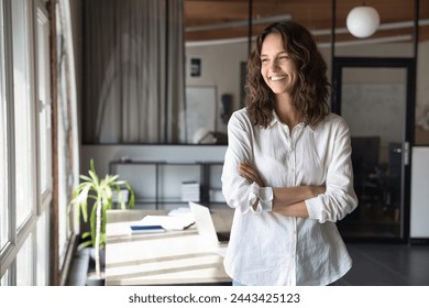 Cheerful young business professional woman in white casual shirt standing in office with arms crossed, looking at window away, smiling, thinking. Happy pensive female manager, entrepreneur portrait - Powered by Shutterstock