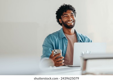 Cheerful young businessman in a casual shirt works with smartphone and laptop in a bright office setting, exuding confidence and professionalism. Modern workplace comfort and productivity. - Powered by Shutterstock