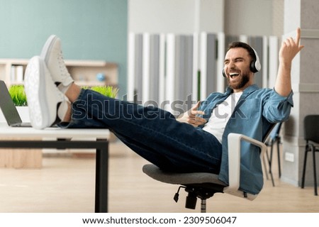 Cheerful young businessman in headphones playing virtual guitar during break in office, happy male employee listening music and putting legs on workdesk, having fun at work, copy space