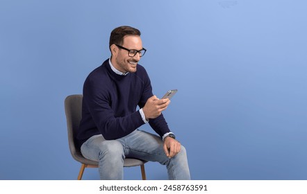 Cheerful young businessman sitting on chair and messaging online over smart phone on blue background - Powered by Shutterstock