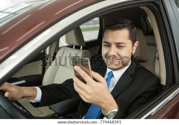 Cheerful young businessman smiling using his smart\
phone while driving technology multitasking danger cars working\
traffic mobility navigation travelling vehicle accident automotive\
luxury