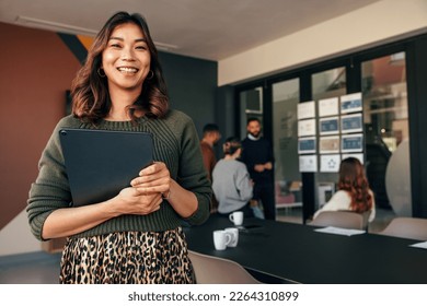 Cheerful young businesswoman smiling at the camera while holding a digital tablet. Happy young businesswoman standing in a boardroom with her colleagues in the background. - Shutterstock ID 2264310899