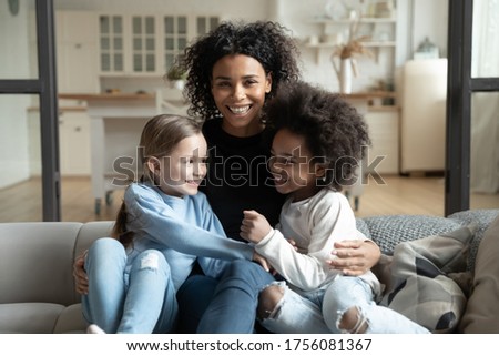 Cheerful young caring African ethnicity single mother embraces multi racial daughters sitting on couch in living room feels overjoyed look at camera. Adoption, childcare, happy family portrait concept
