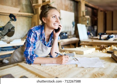 Cheerful young carpenter discussing order details with her customer on smartphone and taking necessary notes, interior of spacious workshop on background