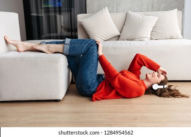 Cheerful Young Casually Dressed Woman Laying On A Floor At Home, Listening To Music With Headphones