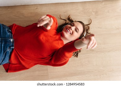 Cheerful Young Casually Dressed Woman Laying On A Floor At Home, Listening To Music With Headphones