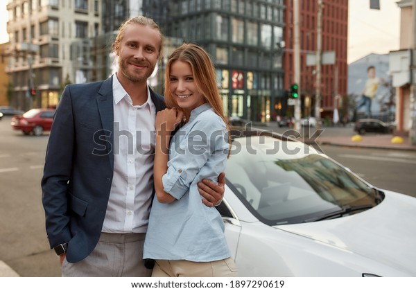 Cheerful young caucasian businessman hugging\
young girl near white car on city street and smiling at camera.\
Lifestyle and success\
concept