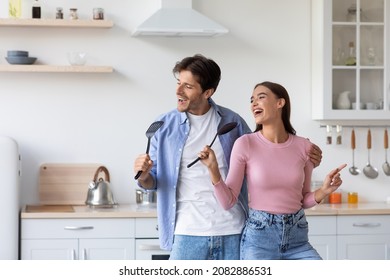 Cheerful young caucasian couple sing in imaginary microphone, have fun and dance in modern kitchen interior. Enjoy cooking together at home during covid-19 pandemic. Positive and facial emotions