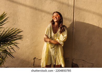 Cheerful young caucasian lady smiles looking toward sun, hugging herself standing near house. Brunette wears sunglasses, dress in summer. Rest time concept - Powered by Shutterstock