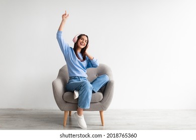 Cheerful young Caucasian lady in wireless headphones sitting in armchair, listening to music, raising her arm, dancing to favorite song, enjoying popular soundtrack, copy space. Home entertainments