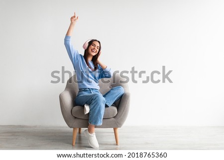 Cheerful young Caucasian lady in wireless headphones sitting in armchair, listening to music, raising her arm, dancing to favorite song, enjoying popular soundtrack, copy space. Home entertainments